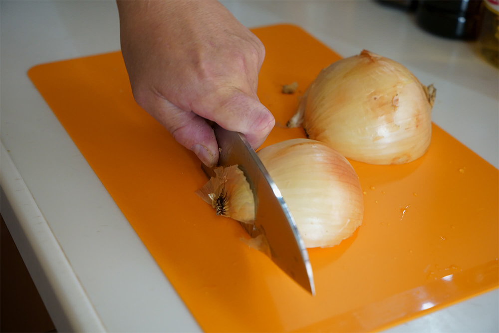 タマネギが転がらないようにまず頭部に包丁を突き刺し、叩きつけるように包丁を下ろして、縦半分に切り分ける。 断面を下にまな板に置き、根元に包丁を刺して押し切る。 左手の平の付け根でタマネギを押さえつけながら、左手指で皮を手前に引きはがして剥く。