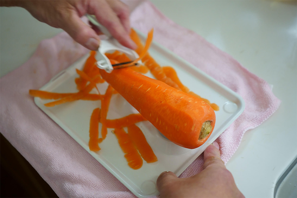 皮剥き器（下参照）を濡れタオルの上に置く。 3本の釘状の固定金具にニンジンを突き刺し、右手で押さえながら、左手でピーラーを使って上半分の皮を剥く。 上下を返して同様に突き刺して反対面も剥く。 ニンジンをまな板に移し、頭部に包丁を突き刺して押し切りした後、目的に応じた切り方で。