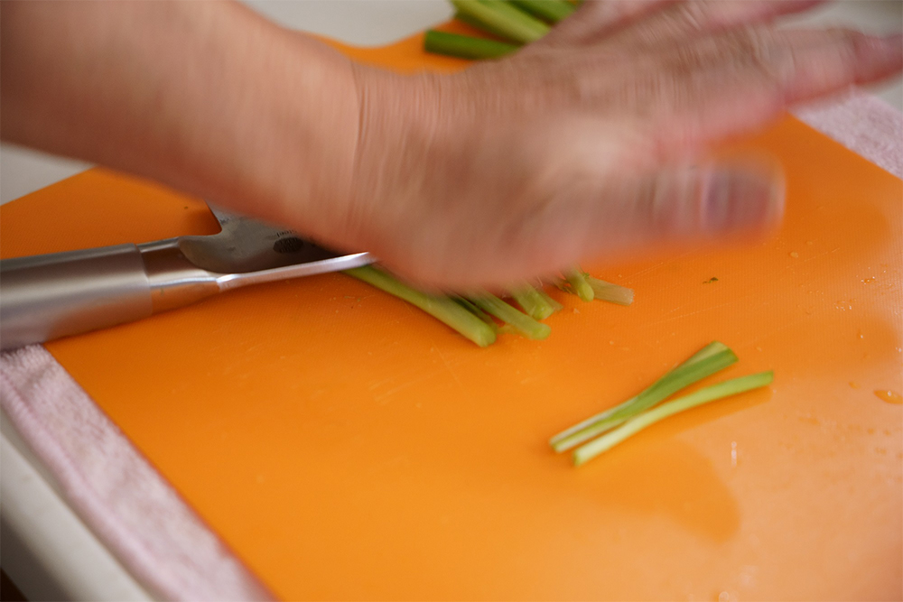 輪ゴムを外して並べ、茎に包丁の面を当てて手の平の付け根で叩き潰す。ニラは根元に近い部分に旨味があるので、その旨味を引き出すように。