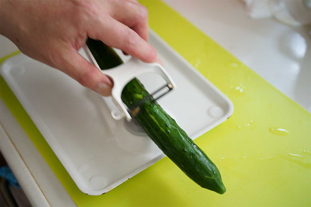 （A）濡れタオルの上に皮むき器を置く。キュウリをくぎ状固定用金具に刺し、ピーラーで皮を一筋だけ剥く。