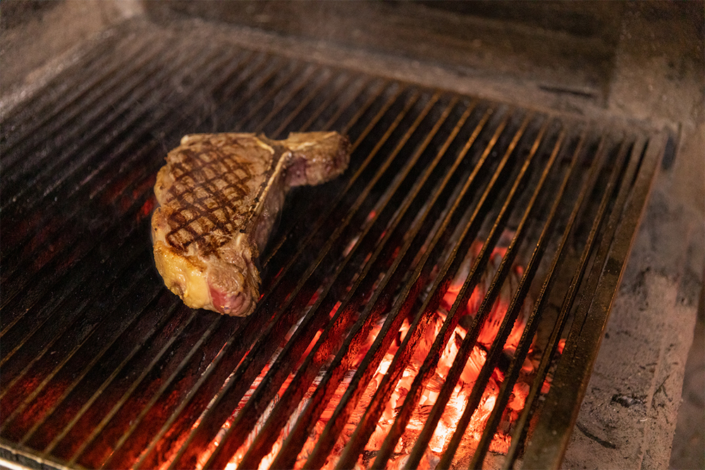 幅３cmのステンレス鋼が0.5cm間隔で連なる焼き網は炉床から高さ10cmの位置に。「強火の近火」で下の熾火から当たる熱だけで肉を焼く。真上のダクトが強力に排気するため、熱はこもらず、放熱性が高い。「輻射熱は要らない」「肉に必要なのは下からの拡散された強い熱」とする渡邊シェフの考え方を実現する構造。