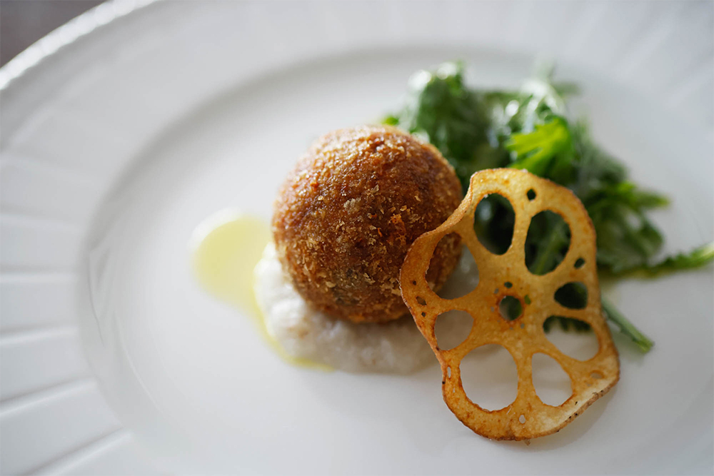 ピュレにしてソースに、角切りにしてアランチーニ（ライスコロッケ）の具材に、素揚げしてチップスに、とレンコンのおいしさを味わい尽くす一皿。「根菜類はオリーブオイルと合わせることで、いろんな表情が出てきます。レンコンは風味が穏やかなので、オリーブオイルもマイルドなポルトガル産を選びました」。仕上げに好みのオイルを回しかけて、味わいの違いを楽しむのもおすすめ。