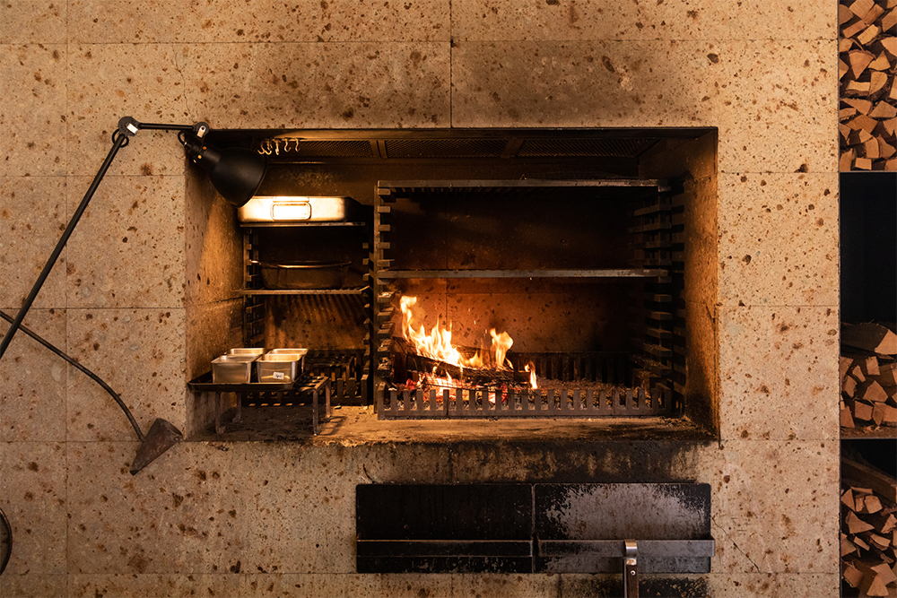 中央で炎を立ち上がらせ、右側で熾火調理、右側一番上で燻製調理、左側は保温と使い分けている。鋼鉄製のフレームにセットする焼き網はのせる段を変えることで火との距離を調節できる。炉の下に灰を受ける引き出しがある。