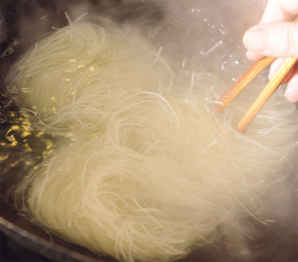 [５]ビーフンを茹でる 中華鍋に鶏ガラスープ450mlを沸かし、塩をひとつまみ加え、ビーフンを入れる。箸でほぐす。（全体で４分弱が目安） POINT：「ビーフン東」では、インスタントを使わず、鶏ガラでとったスープで茹でている！