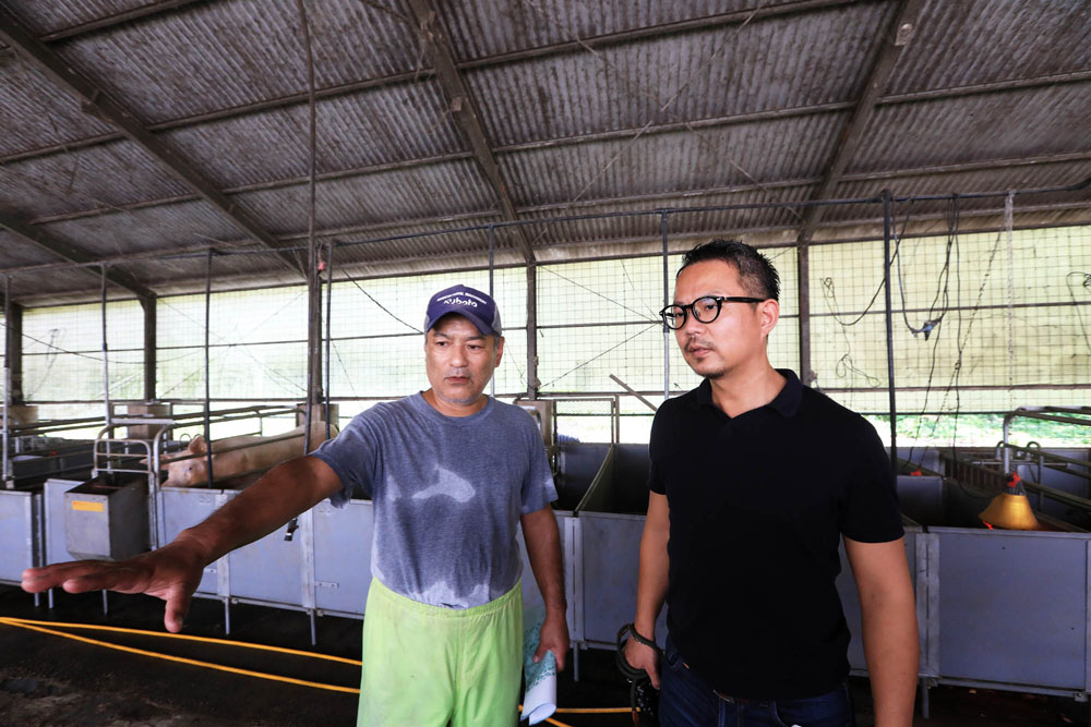 戸外から明るい光が差し込む豚舎で、親川公勇さん（左）の説明に聞き入る久岡シェフ。 「アグー豚といえば“沖縄の黒豚”という漠然とした知識しかなかったけれど。こんなにも時間と手をかけながら、あえて苦労して育てられているんですね」と感慨深げに話す。