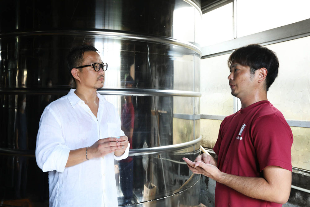 「金武酒造」の奥間尚利さん（右）の説明に熱心に耳を傾ける久岡シェフ。手には、タンク直汲みの新酒が入った利き猪口。金武酒造の泡盛は最低でも１年貯蔵させてから出荷する。
