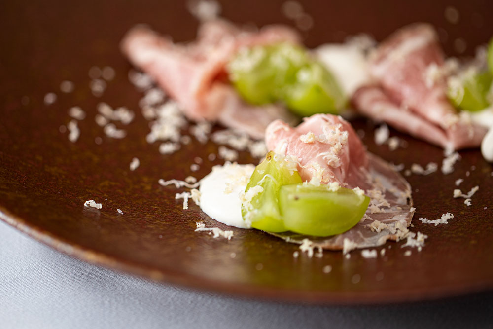 料理：「藁で燻した自家製プロシュートコット」 しっとりした自家製ハムに、クミンとシナモンをほのかに効かせたリコッタチーズと生クリームのソースを添え、シャインマスカット、ホースラディッシュを削りかける。