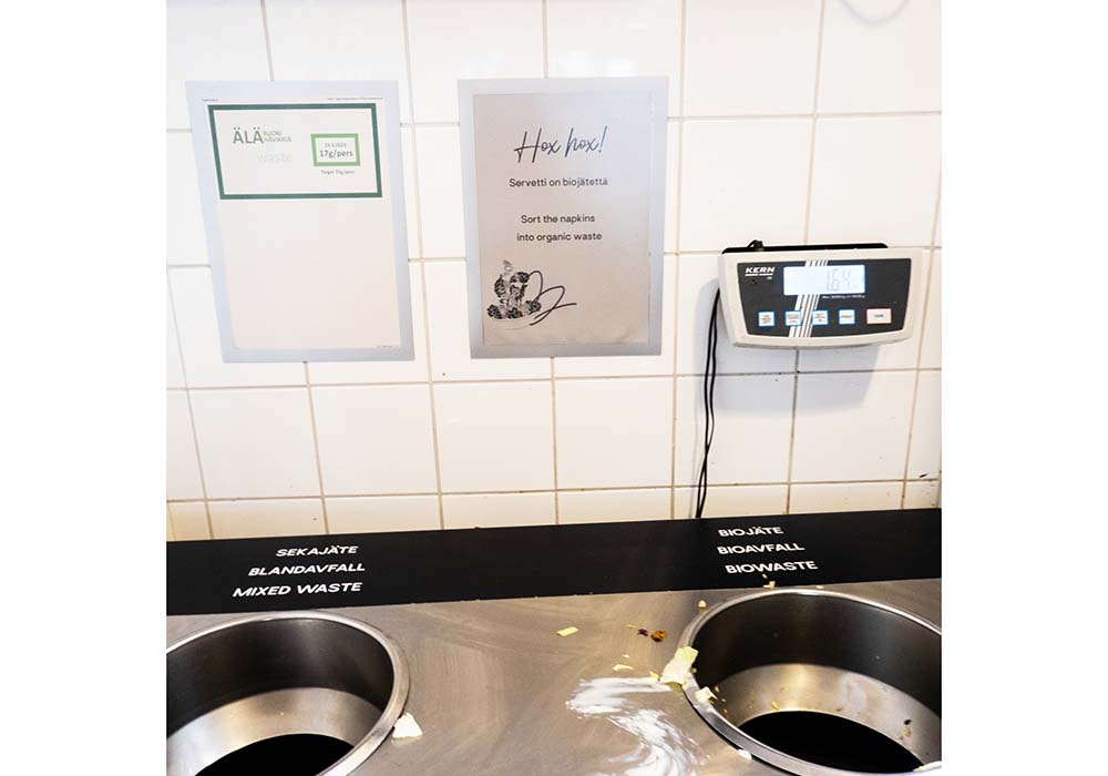 （写真）食べ残しを捨てるゴミ箱と掲示板。食べ残しは１人25gまでが目標。撮影前日の食べ残し量の平均は17gだったと記載されている。