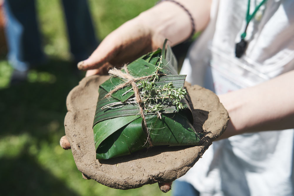 泥包み焼きに挑戦。午前中に採集した泥を捏ねて薄く伸ばし、バナナを無農薬栽培する「木更津ファーム」からもらい受けたバナナの間引き葉にくるんだ鹿肉をタイムやローリエと共に包み込む。