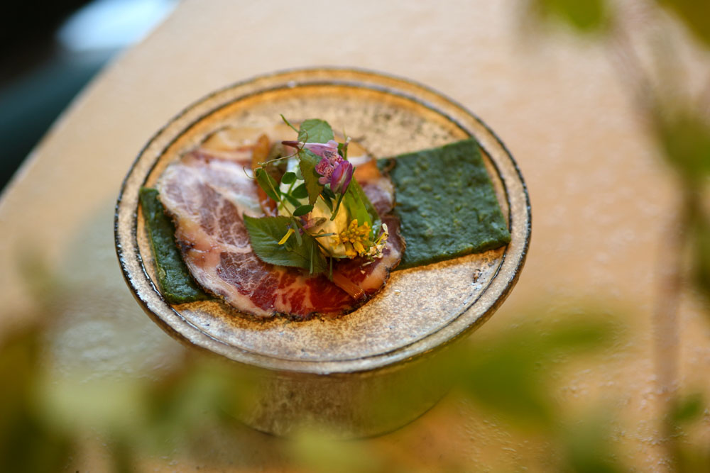 ヨモギのクレープの上に自家製生ハムと地元のチーズ工房「KAWABA CHEESE」のリコッタチーズ。ハナイカダ、ヤブジラミ、タネツケバナ、ハルサキヤマガラシ、カキドオシ、カラスノエンドウ、ウドの葉、ヨモギを添えて。