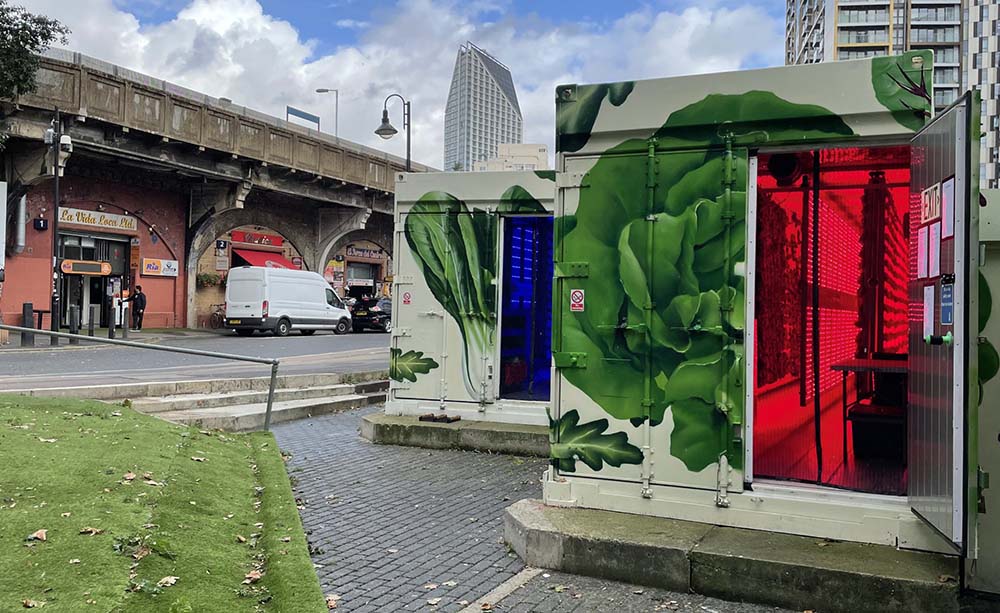 （写真）ロンドン南部、エレファント＆キャッスルの駅近くの公園脇に設置された、２台のコンテナファーム。カラフルで、ヴィヴィッドな野菜のイラストが、通りすがりの人の目を惹く。