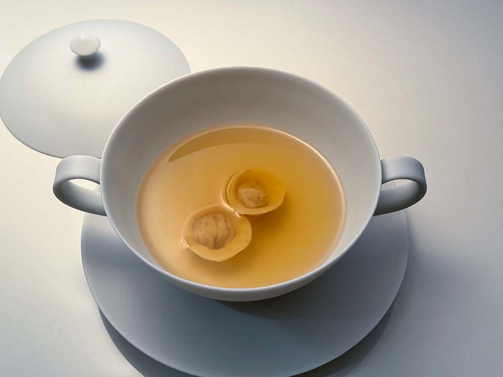 （写真）プレ・ジョーヌ（トウモロコシで育った黄色い身の若鶏）とホロホロ鳥を丸ごと鍋に入れ、36カ月のパルマ産生ハムを加えて、オーブンでじっくり水煮したコンソメスープ。スープの中にはジロール茸と砕いたヘーゼルナッツを詰めたラビオリが浮かぶ。「クリアで品がよく軽く、日本の出汁のようなしみじみとしたおいしさです。シンプルですが味わい深い一皿です」