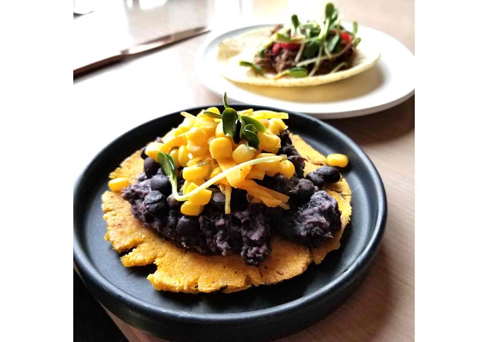 （料理）オワムニの定番「“スリー・シスターズ”のオープンサンド」（16ドル）。潰したトウモロコシのパティにブラックビーンズのピュレをのせ、カボチャのピクルス、トウモロコシの粒をあしらった。これらの野菜はネイティブ・アメリカンたちが畑で並べて栽培したことから“三姉妹”とのニックネームがつけられた。photograph by Gail Irish