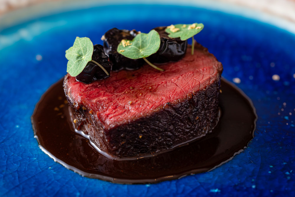 「蝦夷鹿ロースト　伊江島産黒糖キャラメリゼ　イエラムレーズン添え」。フェリーから見た海に浮かぶ伊江島の眺めを思い、今回の旅で購入した青色の皿の中央に盛り付けて。上は伊江島蒸留所のラム酒「イエラム サンタマリア ゴールド」（link ）に漬けた自家製セミドライレーズン。