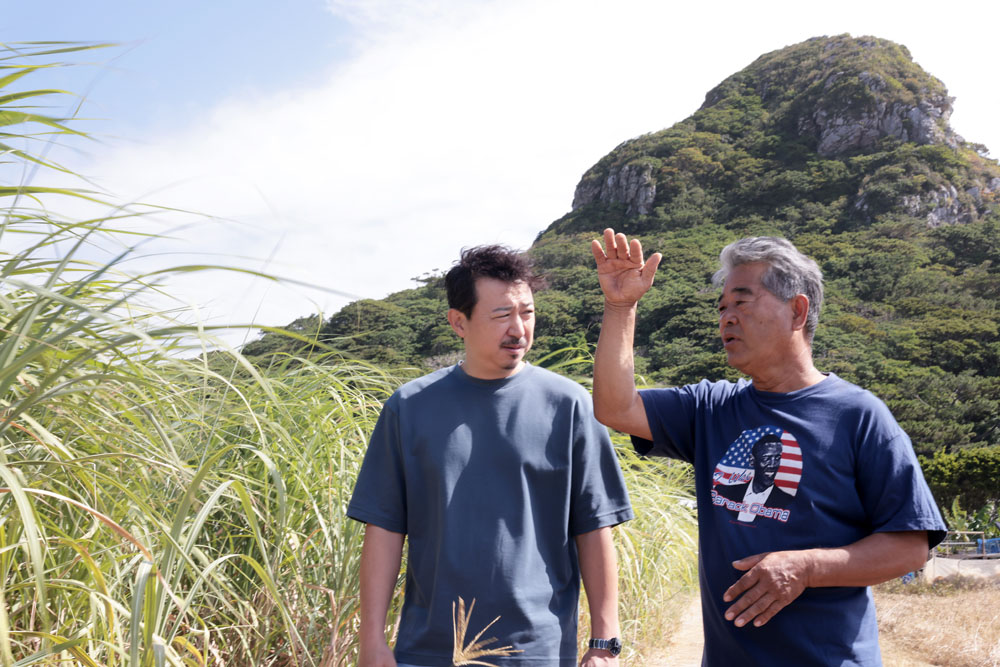 サトウキビ農家の知念和幸さん（右）、宮﨑慎太郎シェフ（左）。後ろにそびえるのは、伊江島のシンボル・城山（ぐすくやま）。