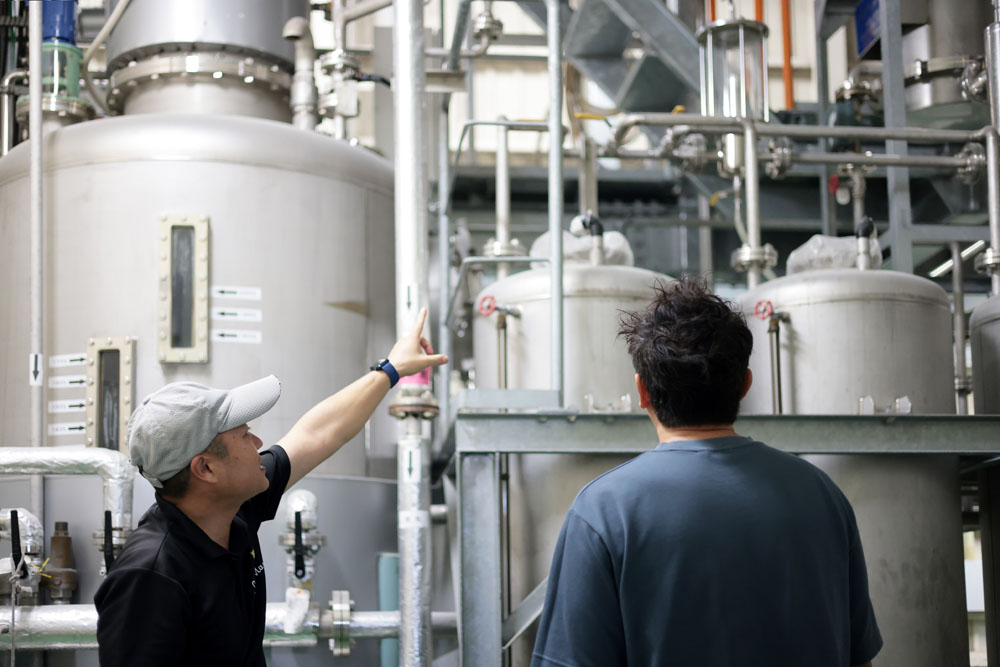 単式蒸留器の説明をする伊江島物産センター 伊江島蒸留所・常務取締役製造責任者の浅香真さん（左）。月桃やジーマミ（落花生）など、素材の香りを移したスパイスドラムを造る際は、黒糖を溶かして発酵させたもろみと一緒に袋に入れた素材を蒸留器に入れ、煮出す。