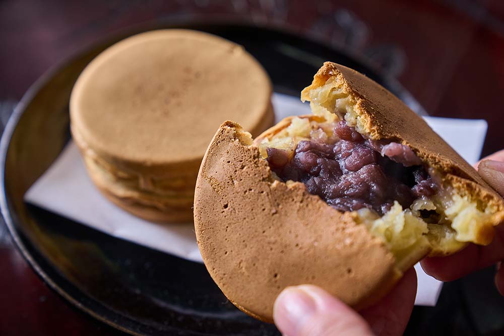 大判焼き（￥120）。表面はサクッと、中はしっとりした生地に、２日前から仕込んだ上品な甘味のあんこがたっぷり。販売期間は、夏場を除く10月〜６月。小豆は北海道産。生地は均一な食感になるように、混ぜ方や材料の調合の仕方を工夫する。「よその味も研究しながら、よそとは違う味で、どうやったらおいしくなるか考えよう」（高橋さん）。せんべいの仕込みは週に１回、日曜にまとめて行っている。