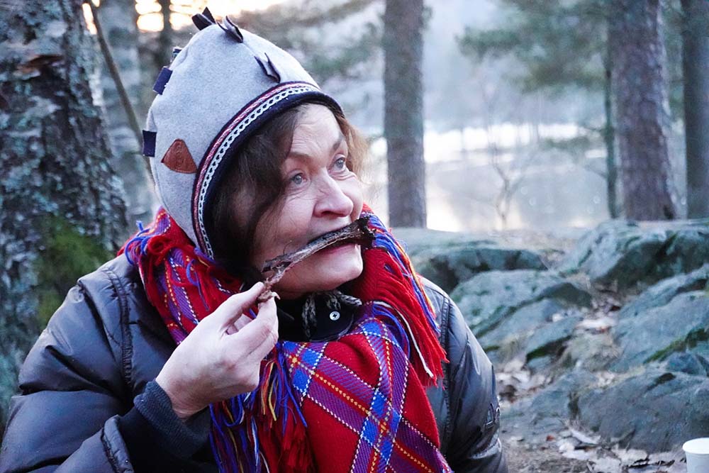（写真）燻製にした後は、さっそく湖の側で焚火をして、肉をさらに焼き上げる。ちょっと焦げたくらいの状態でそのまま食べると、これがまたおいしい。出来上がった肉は自宅で冷凍して数カ月かけて食べる。写真の女性が着用しているのはサーミ人が愛用する伝統的なスカーフだ。