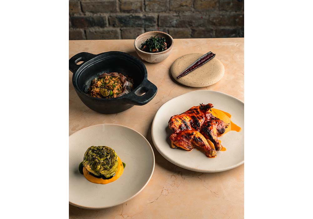 （写真）シグネチャー料理「アカラ」（各種８～10ポンド）以外にも、ビーフ、チキン、ベジ料理など、バランスのとれたアラカルトメニュー（12～34ポンド）が揃う。黒い土鍋の料理は、ココナッツミルクを入れて炊いた店の看板「ジョロフライス」、右「ラゴスチキン」。左手前は、グリルしたキャベツを、”アブヌ・アブヌ”と呼ばれるガーナのグリーンソースと共に。photo by Charlie McKay